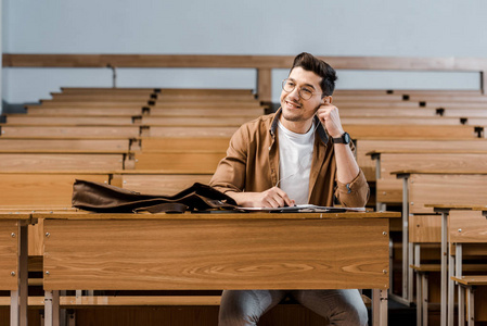 微笑的男学生戴着眼镜坐在桌子上，看着教室