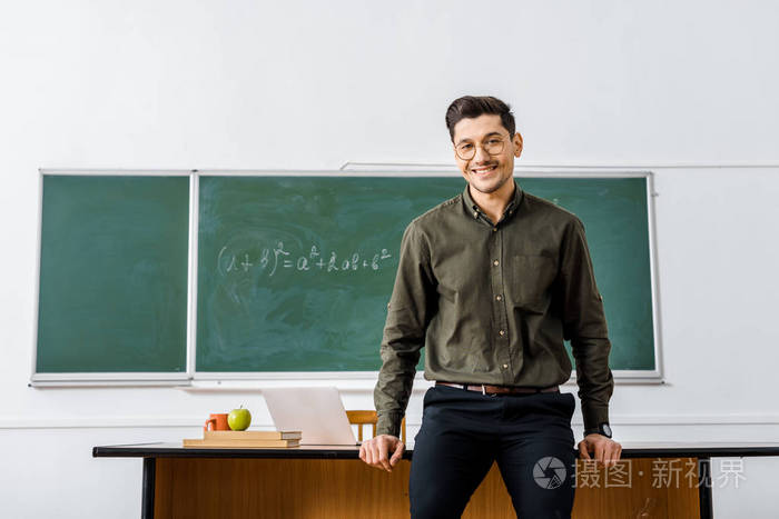 微笑的男老师穿着正式服装看着相机站在教室的桌子旁