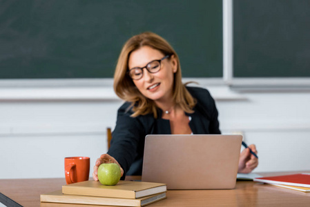 女教师坐在电脑桌旁，在教室里伸手拿苹果