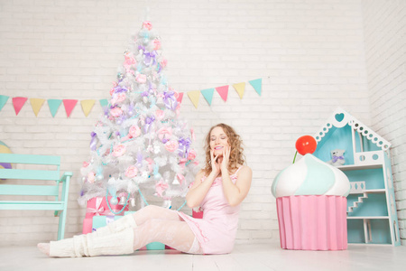 漂亮的年轻女孩穿着睡衣，在她的白色房间里享受着可爱的新年树和巨大的糖果，独自带着礼物。