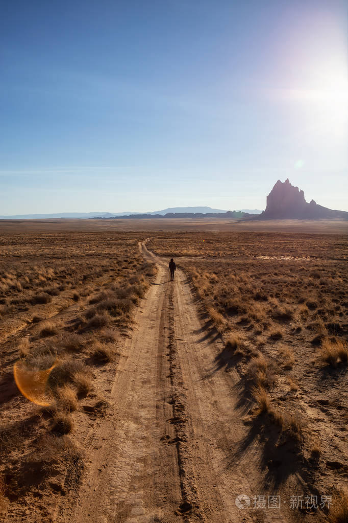 女人走在干燥的沙漠里的土路上，背景是一座山峰。 在新墨西哥州的Shiprock，美国。