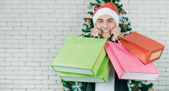 年轻英俊的白种人男子拿着几个大小和颜色的购物袋，幸福和笑脸在大圣诞树前。