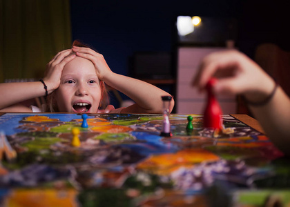 棋盘游戏概念你的行动。 小女孩看了比赛，从动作中震惊。 棋盘游戏领域许多数字。 女孩在家玩棋盘游戏，背景黑暗模糊