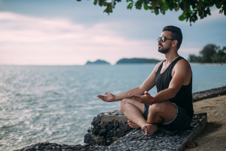 在海边做瑜伽的年轻人。 日落时，一个帅哥坐在海洋上。