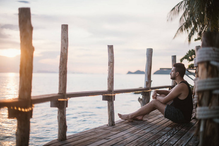 日落时分，一个年轻人坐在一座木桥上。东南亚海边看日落的帅哥