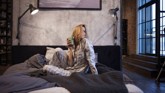 迷人的年轻女子穿着睡衣, 坐在床上喝早茶