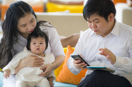年轻漂亮的亚洲华人家庭坐在现代度假村里，工作狂男人在网上用数字平板电脑和手机做生意，而妻子则抱着可爱的小女儿