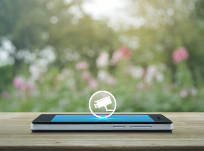 现代智能手机屏幕上的闭路电视摄像机平面图标在木桌上覆盖模糊的粉红色花和树的商业安全在线概念