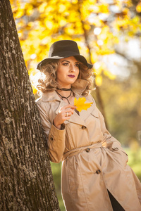 美丽的金发女人，带着奶油外套，长腿和黑色帽子，在秋天的场景中。 一位非常美丽优雅和的女人的肖像，卷发和的腿在秋天公园摆