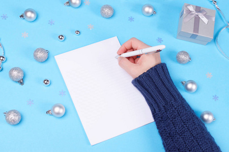 有人在笔记本上写着粉彩蓝色的桌子，上面装饰着圣诞宝，可爱的银礼品盒和雪花纸屑。 新年和圣诞节庆祝概念。