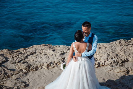 美丽的微笑，年轻的新娘和新郎在海滩上散步，亲吻，在岩石和大海附近举行有趣的婚礼。 塞浦路斯海岸婚礼