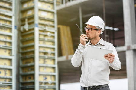 男总工程师戴白色安全安全帽与对讲机和平板检查施工现场。 技术发展和建筑业概念