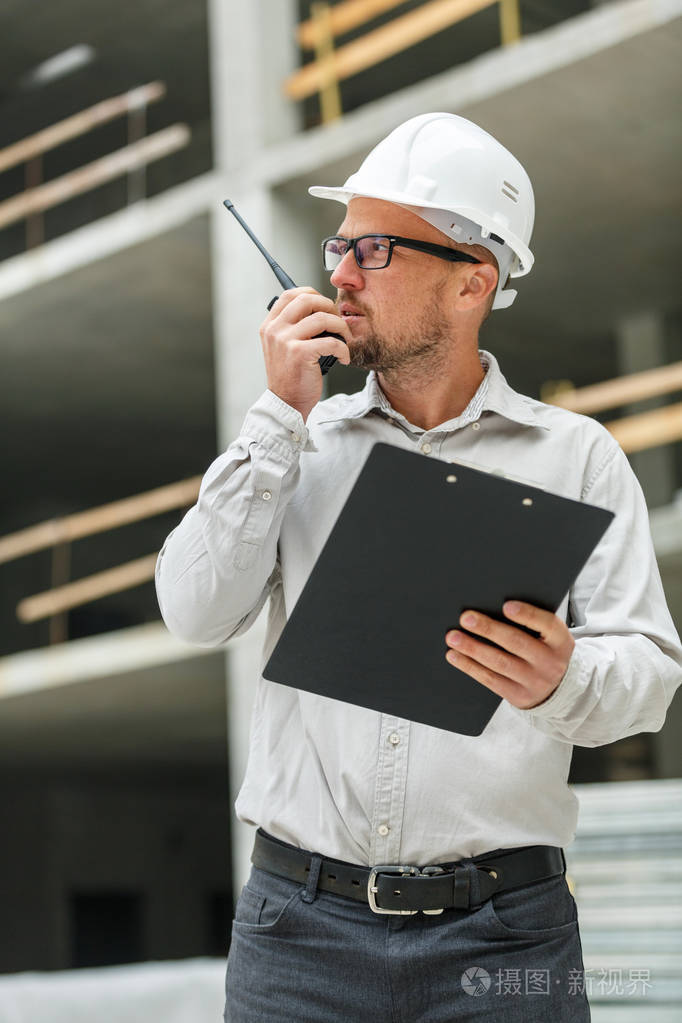男总工程师戴白色安全安全帽与对讲机和剪贴板检查施工现场发展和建筑