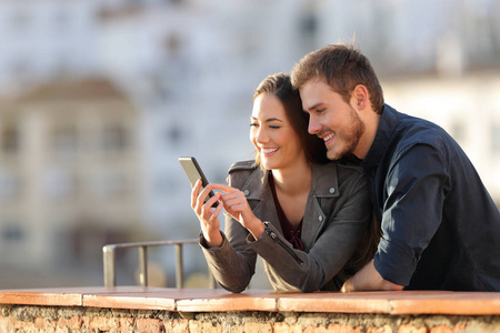 幸福的一对夫妇在日落时在阳台上用智能手机，背景是一个城镇