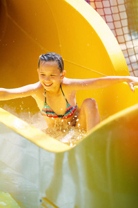 年轻漂亮的女孩穿着条纹五颜六色的泳衣，笑着骑在水上公园的黄色滑梯上。