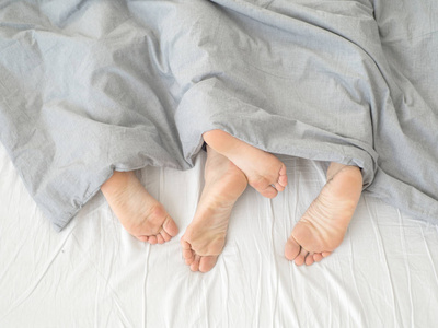双脚并排睡在舒适的床上。 在白色毯子下的床上闭上脚。 一个男人和一个女人光着脚从海湾下面偷看。