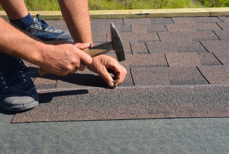 roofer用锤子和钉子在房屋屋顶上安装沥青嵌条.屋顶施工。屋面承包商铺设沥青瓦。