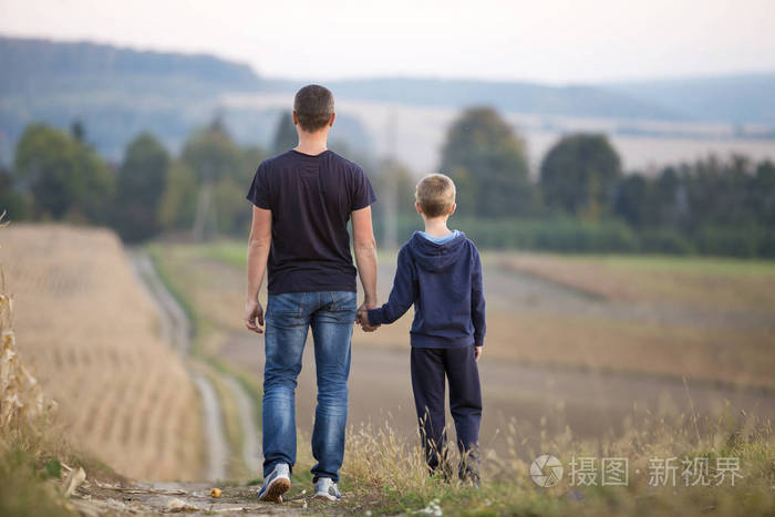 在模糊的雾蒙蒙的绿树和蓝天背景下,年轻的父亲和儿子牵着手在草地上