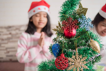 亚洲小女孩带着母亲在圣诞树旁祈祷，为快乐的聚会选择焦点，浅野
