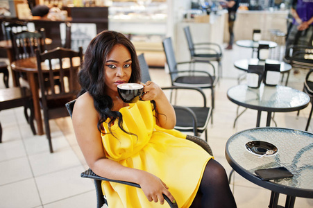 时尚的非裔美国妇女在黄色的黑屋摆姿势在咖啡馆与一杯卡布奇诺。