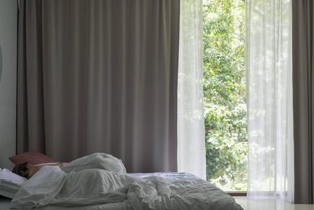 亚洲女人睡在床上，有大窗帘和绿色森林背景。