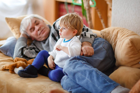 可爱的幼儿男孩和祖父一起看电视节目。小孙子和快乐的退休老人坐在家里, 电视上有卡通片。平静的家庭时间