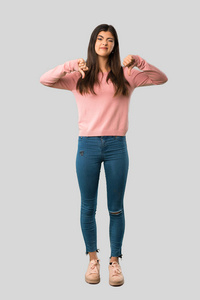 全身的少女，穿着粉红色衬衫，拇指朝下，双手放在孤立的灰色背景上