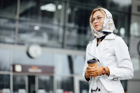 成功的商业女性的肖像，手里拿着一杯热饮料，在去城市街道工作的路上。 办公楼附近有咖啡杯的漂亮女人。