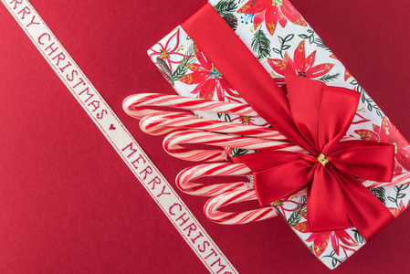 把糖果盒包装在圣诞纸上，上面装饰着红色的蝴蝶结和红色背景礼物上的糖果罐。 贺卡概念。