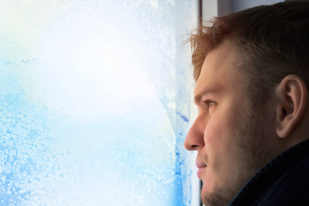 冬天的年轻迷人的男人望着窗外.在白霜和雪花玻璃.