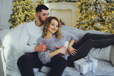 一对相爱的夫妇，坐在圣诞新年树附近的沙发上，彼此温柔地微笑着