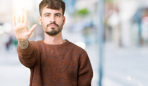年轻的帅哥穿着冬天的毛衣，在孤立的背景上做停止用手掌唱歌。 警告表情，脸上有负面和严肃的手势。