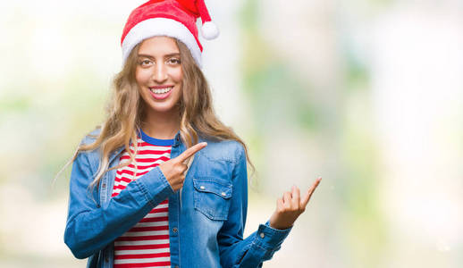 美丽的金发女郎戴着圣诞帽，在孤立的背景上微笑着，看着镜头，双手和手指指向一边。