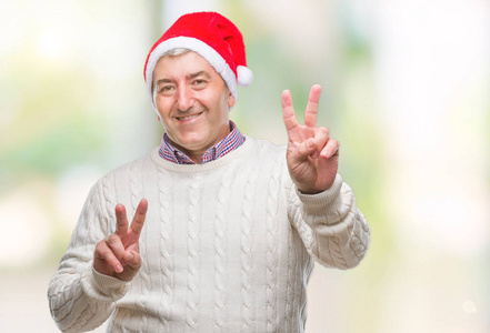 英俊的老人戴着圣诞帽，在孤立的背景上微笑着，看着镜头，手指在做胜利标志。 二号。