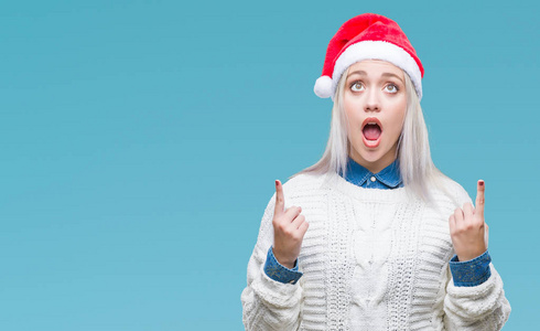年轻的金发女人戴着圣诞帽在孤立的背景上惊讶和惊讶地抬头，用手指和举起的手臂指着。