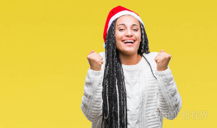 年轻的辫子非洲裔美国女孩戴着圣诞帽，在孤立的背景上庆祝，惊讶和惊讶的成功，举起手臂和睁开眼睛。 赢家概念。