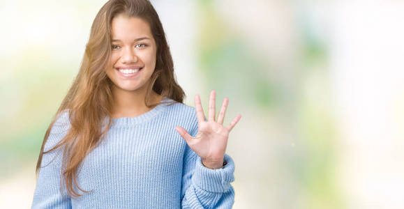 年轻漂亮的黑发女人穿着蓝色的冬季毛衣，在孤立的背景下，用手指举着第五个，微笑着自信和快乐。