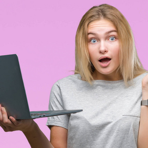 年轻的白种人女人在孤立的背景下使用电脑笔记本电脑，非常高兴和兴奋，胜利者的表情庆祝胜利，微笑着尖叫，举起双手