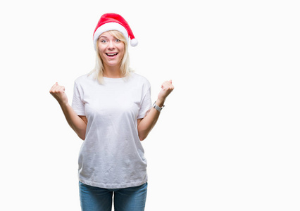 年轻漂亮的金发女人戴着圣诞帽，在孤立的背景上庆祝，惊讶和惊讶的成功，举起手臂和睁开眼睛。赢家概念。
