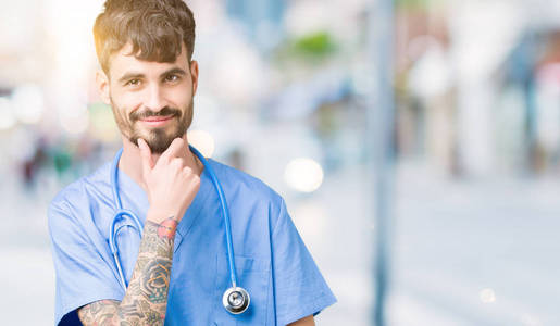 年轻英俊的护士男子穿着外科医生制服，在孤立的背景下，自信地看着相机，微笑着交叉的手臂和举起的手在下巴上。积极思考。