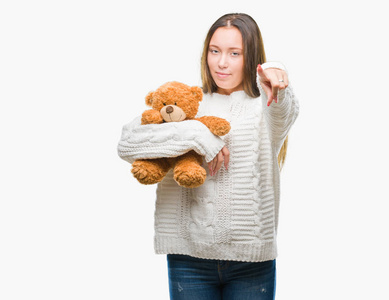 年轻的白种人女人拿着泰迪熊在孤立的背景上指着相机和你，手势，正面积极和自信的手势。
