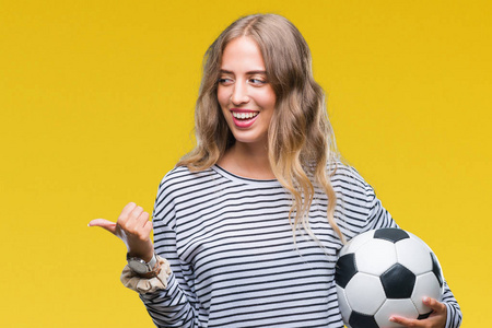 美丽的年轻金发女子，手里拿着足球球，在孤立的背景上指指点点，用用拇指向看，脸上洋溢着幸福的笑容