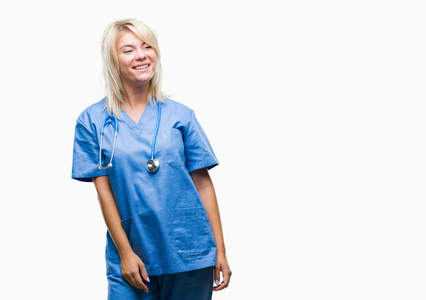 年轻漂亮的金发医生女人穿着医疗制服，在孤立的背景下，看着一边，脸上带着微笑，自然的表情。 自信地笑着。