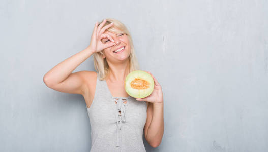 成年白种人女人在灰墙上吃哈密瓜，脸上带着快乐的笑容，用手看着手指，做着确定的标志