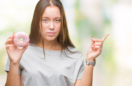 年轻的白种人女人在孤立的背景下吃甜面圈，非常高兴地用手和手指指向一边