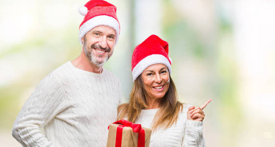 一对中年西班牙裔夫妇戴着圣诞帽，手里拿着礼物，在孤立的背景下，非常高兴地用手和手指指向一边