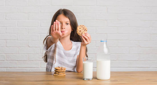 年轻的西班牙裔儿童坐在桌子上喝牛奶，张开手吃巧克力，做停止标志，严肃而自信的表情，防御手势