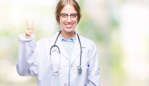 美丽的年轻金发医生女人穿着医疗制服，在孤立的背景上显示和手指第二，同时微笑自信和快乐。