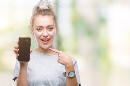 年轻的金发女人在孤立的背景上显示智能手机屏幕，非常高兴地用手和手指指着。