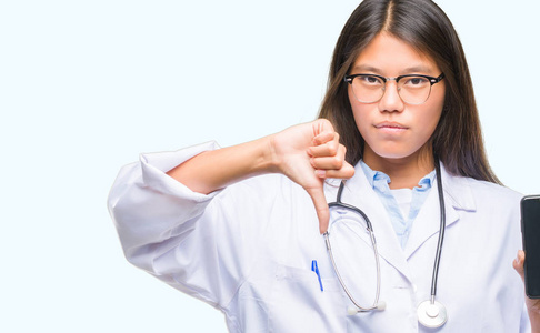 年轻的亚洲医生女人拿着智能手机在孤立的背景上，愤怒的脸，消极的迹象，显示不喜欢，拇指向下拒绝概念